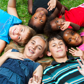 Auf dem Bild liegen sechs Kinder auf dem Rasen in einem Kreis und lächeln in die Kamera. Sie haben ihre Köpfe nah beieinander, sodass sie einen geschlossenen Kreis bilden. Die Kinder tragen bunte Sommerkleidung und scheinen fröhlich und entspannt zu sein. Einige von ihnen winken der Kamera zu. Der Hintergrund besteht aus grünem Gras.