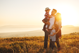 Eine Familie steht auf einer Wiese bei Sonnenuntergang. Ein Mann hält ein kleines Kind im Arm, während eine Frau und ein weiteres Kind neben ihnen stehen. Alle lächeln. Im Hintergrund erstreckt sich eine Landschaft mit Hügeln und einem Gewässer.