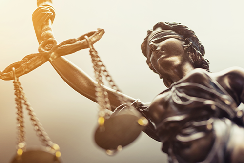 Das Bild zeigt eine Nahaufnahme einer Statue der Justitia, der römischen Göttin der Gerechtigkeit. Die Figur ist mit verbundenen Augen dargestellt, was für Unparteilichkeit steht. In ihrer rechten Hand hält sie eine Waage. Die Statue ist in einem bronzenen Farbton gehalten.