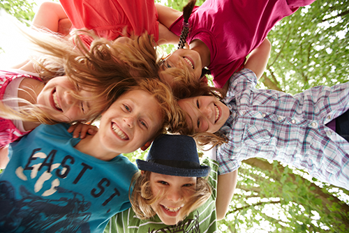 Auf dem Bild sind sechs fröhliche Kinder zu sehen, die in einem Kreis stehen und ihre Köpfe zusammenstecken. Sie lachen und schauen nach unten in die Kamera. Die Kinder tragen bunte Kleidung. Im Hintergrund sind grüne Blätter von Bäumen zu erkennen.
