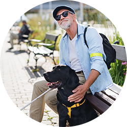 Ein Mann sitzt auf einer Parkbank aus Holz am Gehwegrand. Der Mann hat eine Sonnenbrille auf und hält einen Blindenstock in der Hand. Vor ihm sitzt ein schwarzer Hund. Der Mann guckt nach oben.
